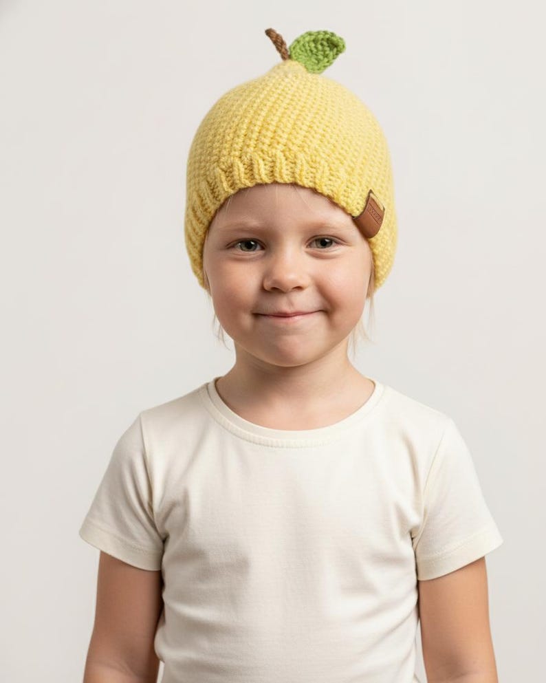 May include: A bright yellow crochet beanie hat shaped like a pear, featuring a small brown stem and a single green leaf on top. The hat has a ribbed brim and a small brown rectangular tag attached to the side, worn over a plain white t-shirt.