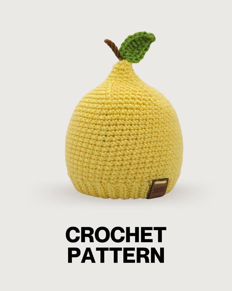 May include: A yellow crochet hat shaped like a pear with a brown stem and a green leaf on top. The hat features a textured stitch pattern and a small brown rectangular tag with the text JULIVIA attached to the bottom edge. The text CROCHET PATTERN is below.