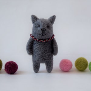 Pu&ograve; includere: Un giocattolo di gatto in feltro grigio con un collare a righe rosse e bianche. Il gatto &egrave; in piedi su una superficie bianca con cinque palline di feltro colorate.