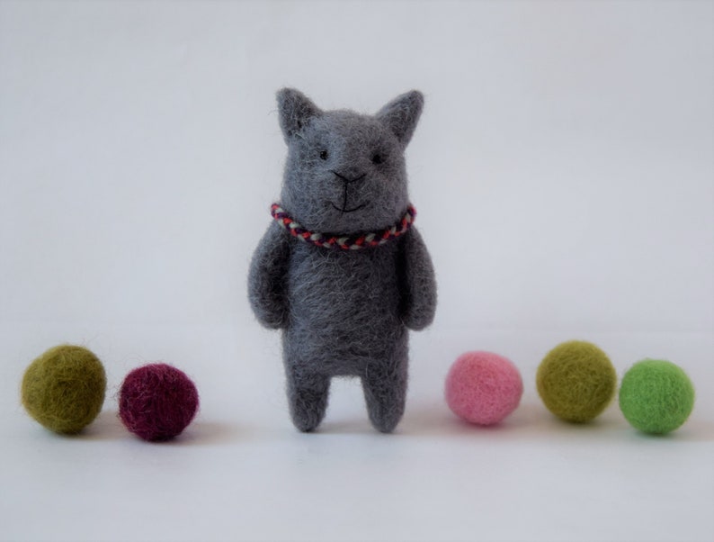 Pu&ograve; includere: Un giocattolo di gatto in feltro grigio con un collare a righe rosse e bianche. Il gatto &egrave; in piedi su una superficie bianca con cinque palline di feltro colorate.