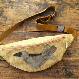 Peut inclure: Une banane en tissu beige avec une fermeture à glissière et une applique d'oiseau marron. La banane a une sangle réglable marron.