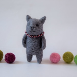 Pu&ograve; includere: Un giocattolo di gatto in feltro grigio con un collare di corda rosso e bianco. Il gatto &egrave; in piedi su una superficie bianca con cinque palline di feltro colorate.
