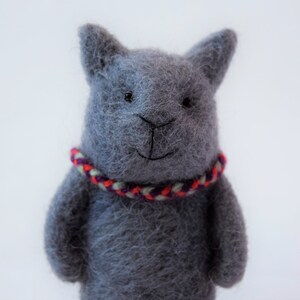 Pu&ograve; includere: Un gatto di peluche grigio in feltro con un collare intrecciato rosso, bianco e viola.