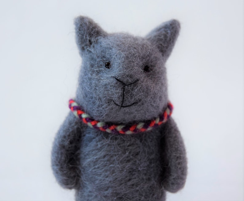 Pu&ograve; includere: Un gatto di peluche grigio in feltro con un collare intrecciato rosso, bianco e viola.