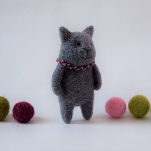 Pu&ograve; includere: Un giocattolo di gatto in feltro grigio con un collare a righe rosso e bianco. Il gatto &egrave; in piedi su una superficie bianca con cinque palline di feltro colorate di fronte a lui.