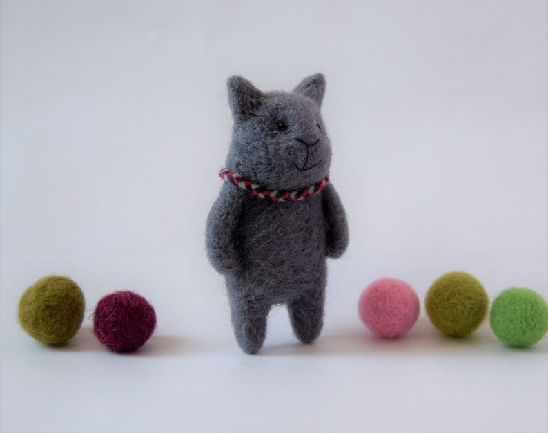 Pu&ograve; includere: Un giocattolo di gatto in feltro grigio con un collare a righe rosso e bianco. Il gatto &egrave; in piedi su una superficie bianca con cinque palline di feltro colorate di fronte a lui.