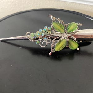 May include: Silver-toned hair clip with a butterfly and floral design. The butterfly has green, oval-shaped accents. The clip also features a cluster of iridescent, round gems. The clip is approximately 4 inches long.