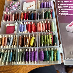May include: A clear plastic storage container filled with numerous spools of embroidery floss in a wide variety of colors. The container features multiple compartments, and the adjacent packaging displays the text FLOSS C and Keep Floss Cle.