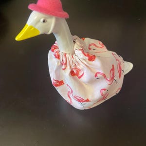 May include: A white goose figurine with a yellow beak and a pink hat. The goose is wearing a pink and white fabric dress with a flamingo pattern. The figurine is on a dark surface.
