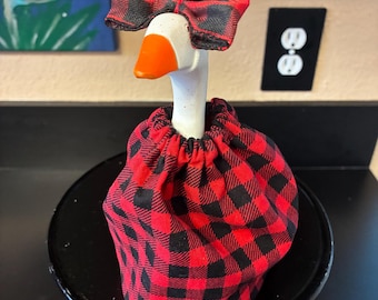 May include: A white goose figurine with an orange beak, wearing a red and black buffalo plaid fabric dress and a matching red and black buffalo plaid bow attached to the top of its head. The goose is positioned on a black circular stand.
