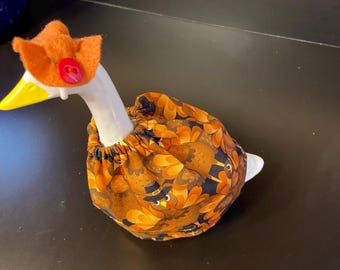 May include: A ceramic goose figurine with a white neck and head, a yellow beak, and a festive orange hat with a red button. The goose is wearing a fabric dress with a repeating pattern of turkeys in shades of orange and brown.