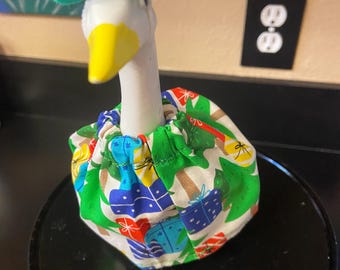 May include: A ceramic goose figurine wearing a green hat and a festive Christmas-themed collar. The collar features a pattern of green Christmas trees and colorful gift boxes on a white background. The goose has a yellow beak.