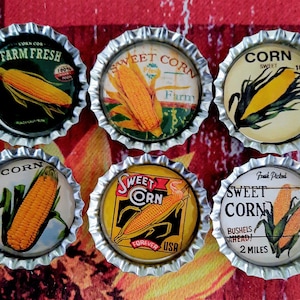 May include: Six vintage-style bottle caps with corn illustrations. The caps feature text like "Farm Fresh" and "Sweet Corn," with images of corn on the cob. The caps have a silver, crimped edge and are arranged on a red and orange patterned surface.