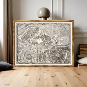 Puede incluir: Una pieza de arte mural rectangular enmarcada que presenta un mapa histórico detallado en blanco y negro de una ciudad, incluyendo un río, trazados de calles urbanas y estructuras fortificadas, en un marco de madera natural.