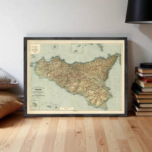 Pode incluir: Mapa vintage emoldurado da Sicília, Itália, em uma moldura de madeira escura. O mapa apresenta informações geográficas detalhadas com um esquema de cores bege e marrom, em um fundo azul claro. O mapa é exibido em um piso de madeira.