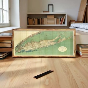 Peut inclure: Une carte vintage encadrée de Long Island, New York, avec un cadre en bois et des tons verts et beiges. La carte est posée sur un sol en bois clair. Des crayons sont au premier plan et des livres sont empilés sur le sol et l'étagère.