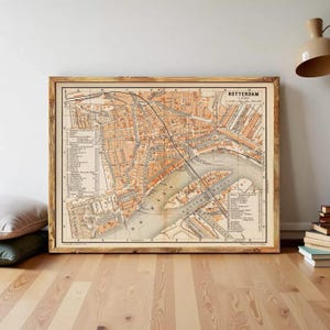 Puede incluir: Un mapa vintage enmarcado de Rotterdam, Países Bajos, en tonos naranja, marrón y beige. El mapa detalla el diseño de la ciudad, incluyendo calles, vías fluviales y puntos de interés etiquetados. El mapa se muestra en un marco de madera.