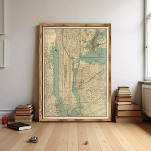 May include: A framed vintage map of New York City, featuring a detailed street layout with red lines indicating subway routes. The map has a sepia tone, framed in a light brown wooden frame. The map is leaning against a wall, with books and small toys on the floor.