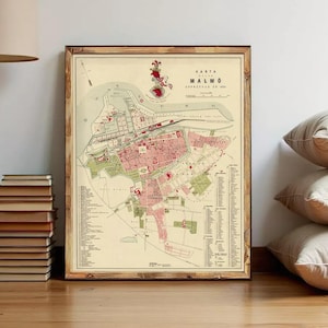 May include: Framed vintage map of Malmö, Sweden, featuring a detailed city plan in red and green hues. The map, titled "Karta Malmö," is encased in a wooden frame and leans against a wall. A stack of books and pillows are in the foreground.