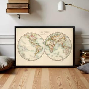 May include: Framed vintage world map print. The map is divided into two hemispheres, with detailed illustrations of continents and countries. The map is in a black frame and is displayed on a wooden floor. The text "THE WORLD HEMISPHERES" is printed at the top.