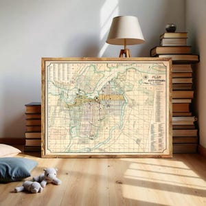 Op de afbeelding: Een ingelijste vintage kaart getiteld Plan of the City of Ottawa, met een raster van straten, rivierlopen en topografische details in gedempte beige, groene en gele tinten, gevat in een eenvoudige houten lijst met een windroos.