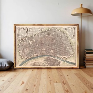 Puede incluir: Una impresión de mapa de estilo antiguo enmarcada que presenta una vista detallada a vista de pájaro de un plano de ciudad con un río sinuoso, cuadrículas de calles intrincadas y elementos cartográficos decorativos en un marco de madera clara.