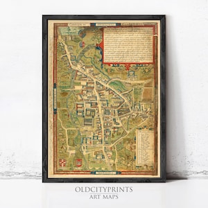 May include: A framed historical map of Cambridge, England, featuring detailed illustrations of city streets, buildings, and green spaces. The map includes Latin text, a numbered legend, and heraldic shields, all presented in a dark wooden frame.