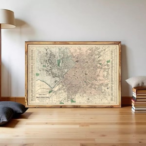 Op de afbeelding: Een ingelijste vintage kaart van Manchester, Engeland, in een houten frame. De kaart toont gedetailleerde stratenplannen, groene ruimtes en de tekst "Jacobs Manchester". De kaart is in beige, groene en bruine tinten.