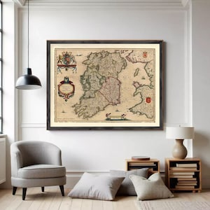 Op de afbeelding: Een ingelijste antieke kaart van Ierland, met gedetailleerde cartografie en historische illustraties. De kaart staat in een donkere houten lijst, weergegeven op een witte muur boven een grijze fauteuil en kussens. De kleuren van de kaart zijn beige, groen en rood.