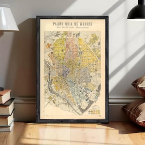 Op de afbeelding: Een ingelijste vintage kaart met de titel PLANO GUIA DE MADRID, BAILLY-BAILLIERE E HIJOS - Editores propietarios. De kaart toont een gedetailleerd stratenplan van Madrid in tinten geel, blauw en beige, in een donkere, getextureerde houten lijst.