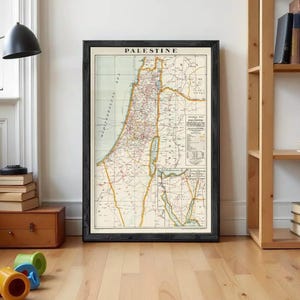 May include: A framed vintage map of Palestine featuring a cream-coloured background with black text, thin red and yellow lines delineating borders and routes, and a detailed legend in the lower right corner, all enclosed in a dark wooden frame.
