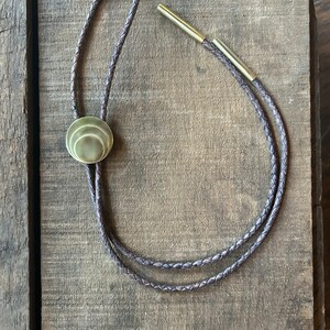 May include: A brown leather bolo tie with a gold-toned metal tip and a round, layered, gold-toned metal pendant.