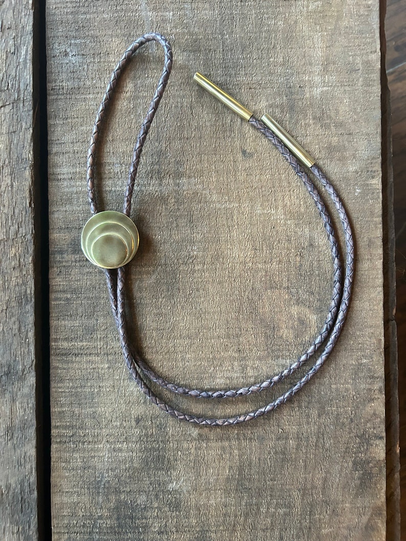 May include: A brown leather bolo tie with a gold-toned metal tip and a round, layered, gold-toned metal pendant.