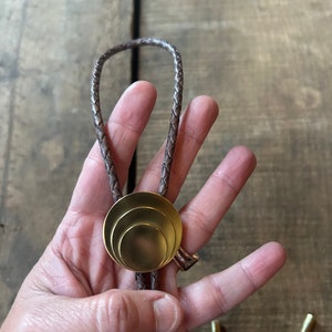 May include: A brown leather bolo tie with a gold-toned, concentric circle pendant.