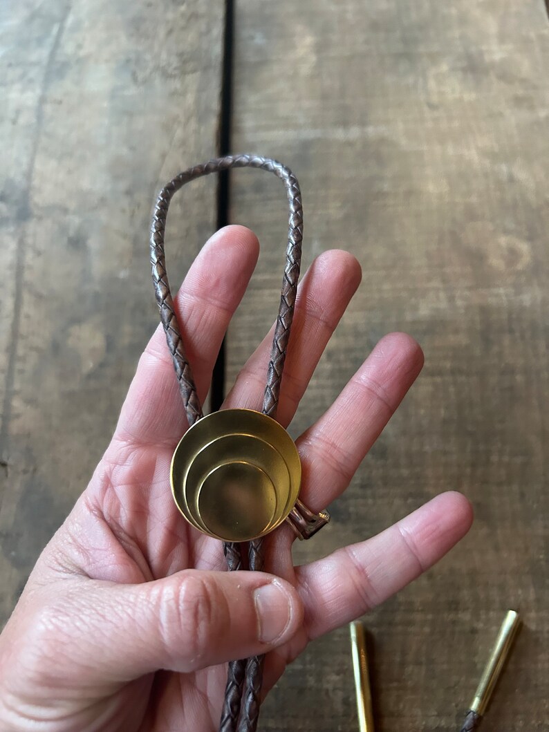 May include: A brown leather bolo tie with a gold-toned, concentric circle pendant.