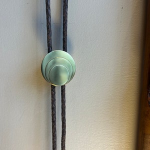 May include: A brown leather bolo tie with a circular, brass, concentric circle design.