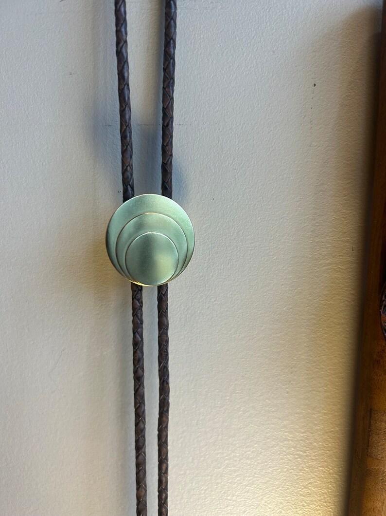 May include: A brown leather bolo tie with a circular, brass, concentric circle design.