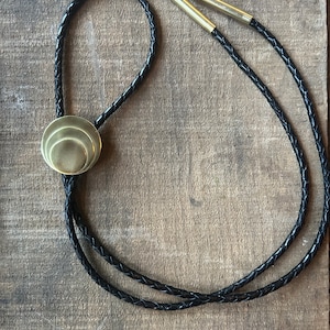 May include: A black braided leather bolo tie with a gold-toned, layered circular pendant.