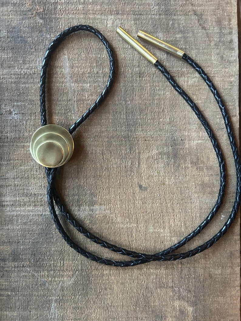 May include: A black braided leather bolo tie with a gold-toned, layered circular pendant.
