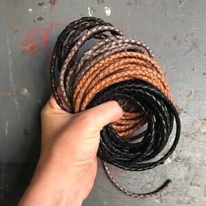 May include: A close-up of a hand holding a stack of braided leather cords in black, brown, and gray.
