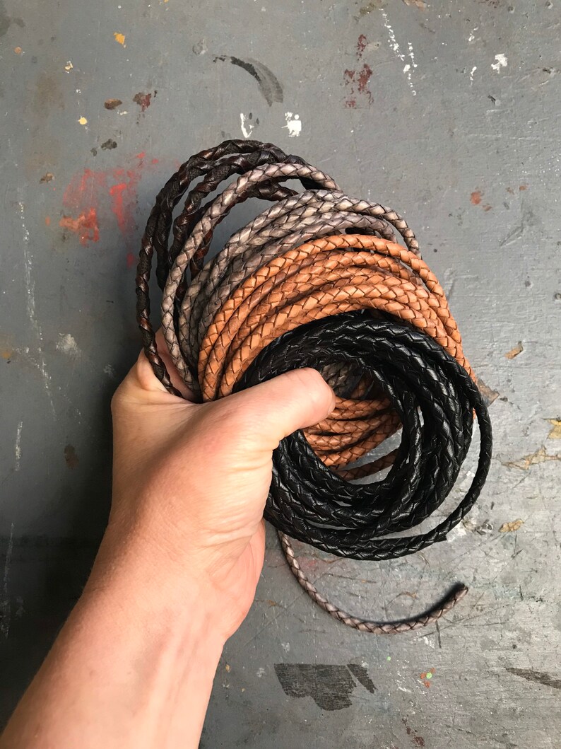 May include: A close-up of a hand holding a stack of braided leather cords in black, brown, and gray.