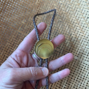 May include: A brown leather bolo tie with a gold-toned circular pendant featuring three concentric circles.