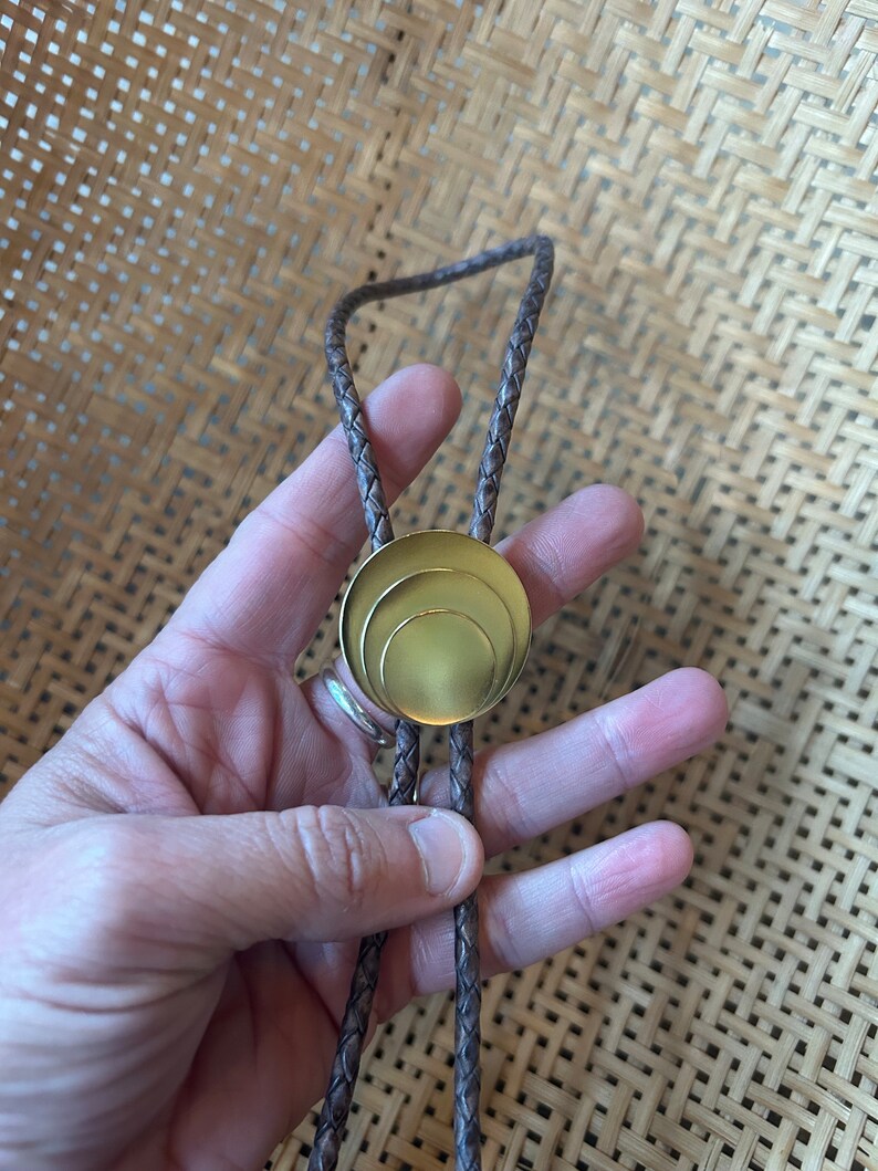 May include: A brown leather bolo tie with a gold-toned circular pendant featuring three concentric circles.