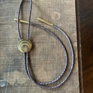 May include: A brown leather bolo tie with a gold-toned metal tip. The tip is a layered circle design.