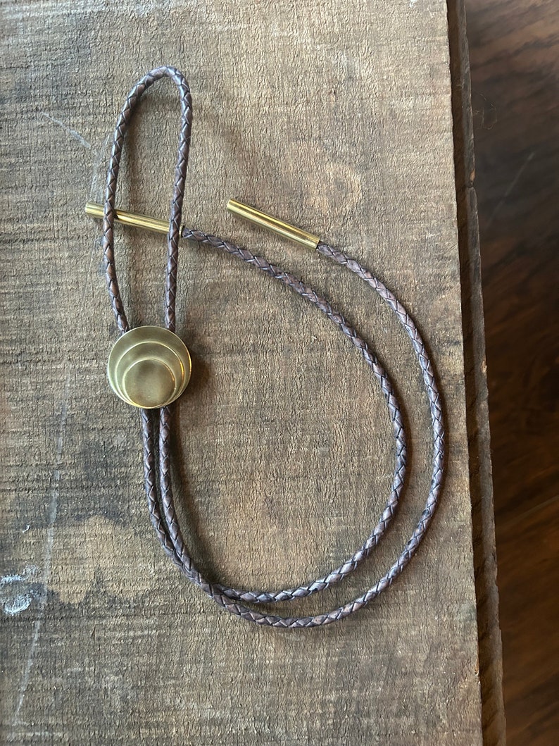 May include: A brown leather bolo tie with a gold-toned metal tip. The tip is a layered circle design.