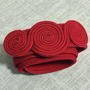 May include: A bright red bracelet constructed from thick, braided cord. The design features three prominent, circular spiral motifs arranged horizontally across the top, connected to a flexible, coiled band that wraps around the wrist.