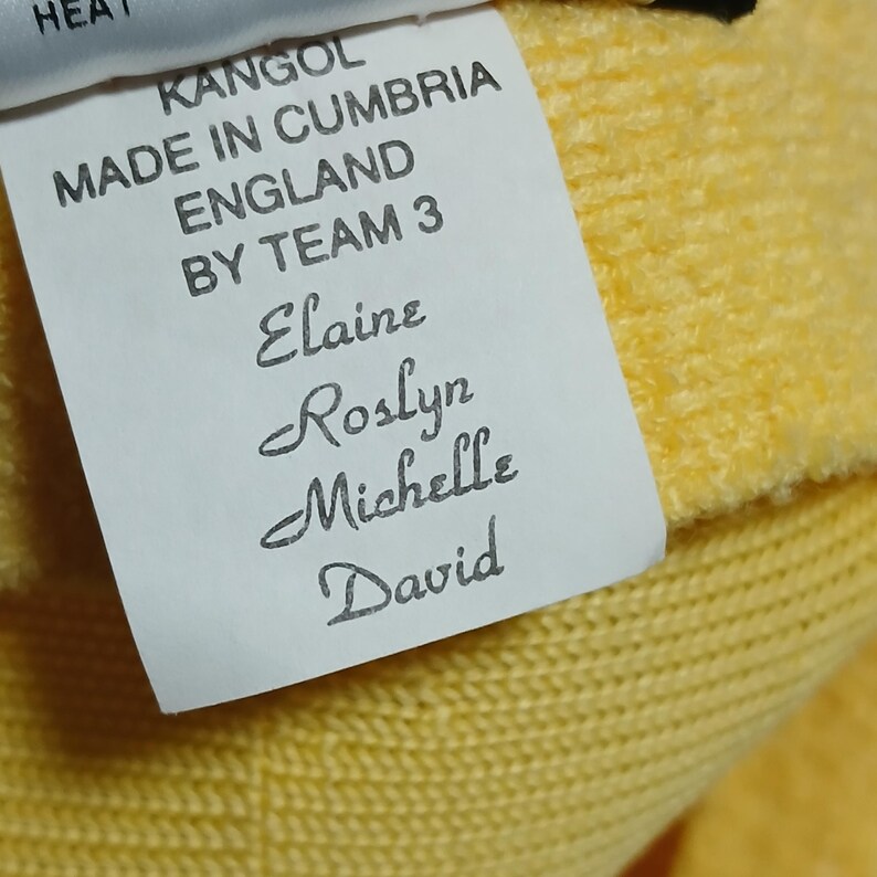 Puede incluir: Primer plano de una etiqueta con el texto "KANGOL MADE IN CUMBRIA ENGLAND BY TEAM 3". Debajo del texto est&aacute;n los nombres Elaine, Roslyn, Michelle y David. La etiqueta est&aacute; sobre una tela amarilla tejida.