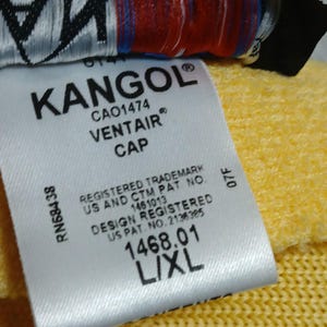 Puede incluir: Primer plano de una etiqueta blanca con el logotipo KANGOL y el texto "VENTAIR CAP". La etiqueta tambi&eacute;n incluye la talla "L/XL" e informaci&oacute;n de registro. El fondo es amarillo.