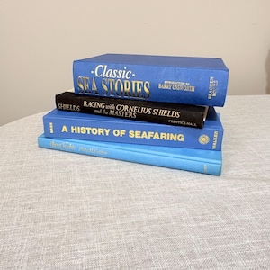 May include: A stack of four hardcover books with nautical themes. The top book is blue with gold text reading Classic Sea Stories. Below it is a black book titled Racing with Cornelius Shields and the Masters, a blue book titled A History of Seafaring, and a light blue book titled Great Yachts.