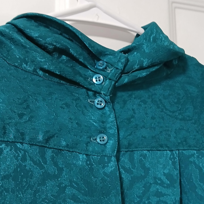 Peut inclure: Chemisier de couleur teal avec un col montant et trois boutons assortis. Le tissu pr&eacute;sente un motif floral subtil en relief. Le chemisier est suspendu &agrave; un cintre blanc sur fond blanc.